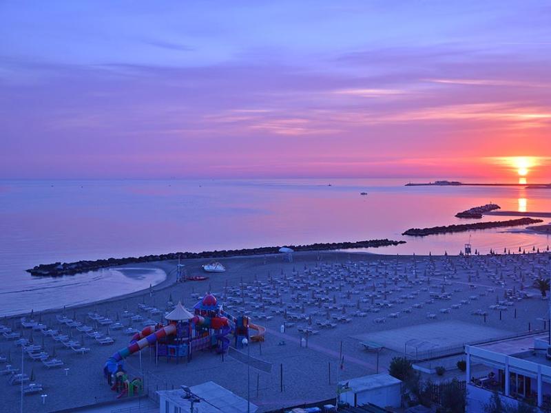 Tramonto sulla spiaggia con ombrelloni e parco giochi, cielo colorato di viola e arancio.